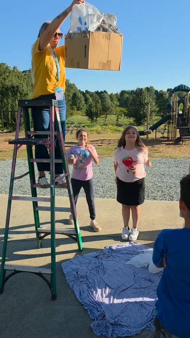 A teacher on a ladder holding up a box for an outdoor experiment as students watch